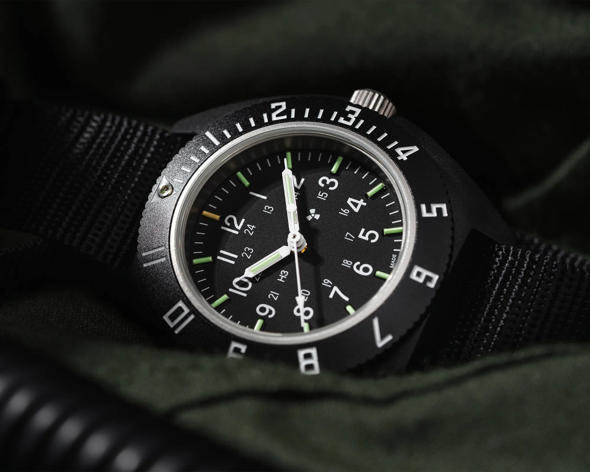 Black tactical wristwatch with white numerals, green luminous markers, and a black fabric strap on dark fabric.