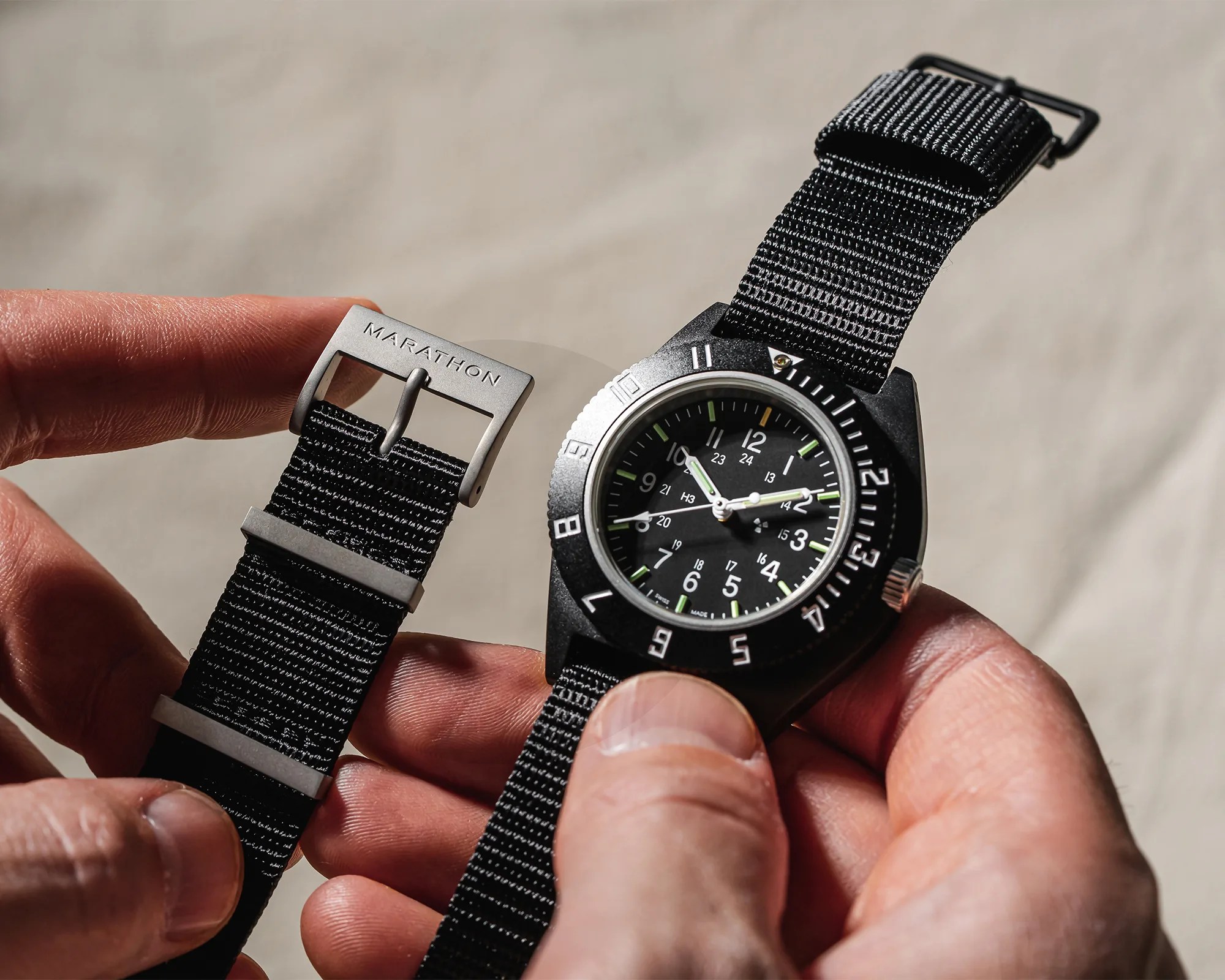 Black Marathon wristwatch with white numerals and black nylon strap held in two hands.
