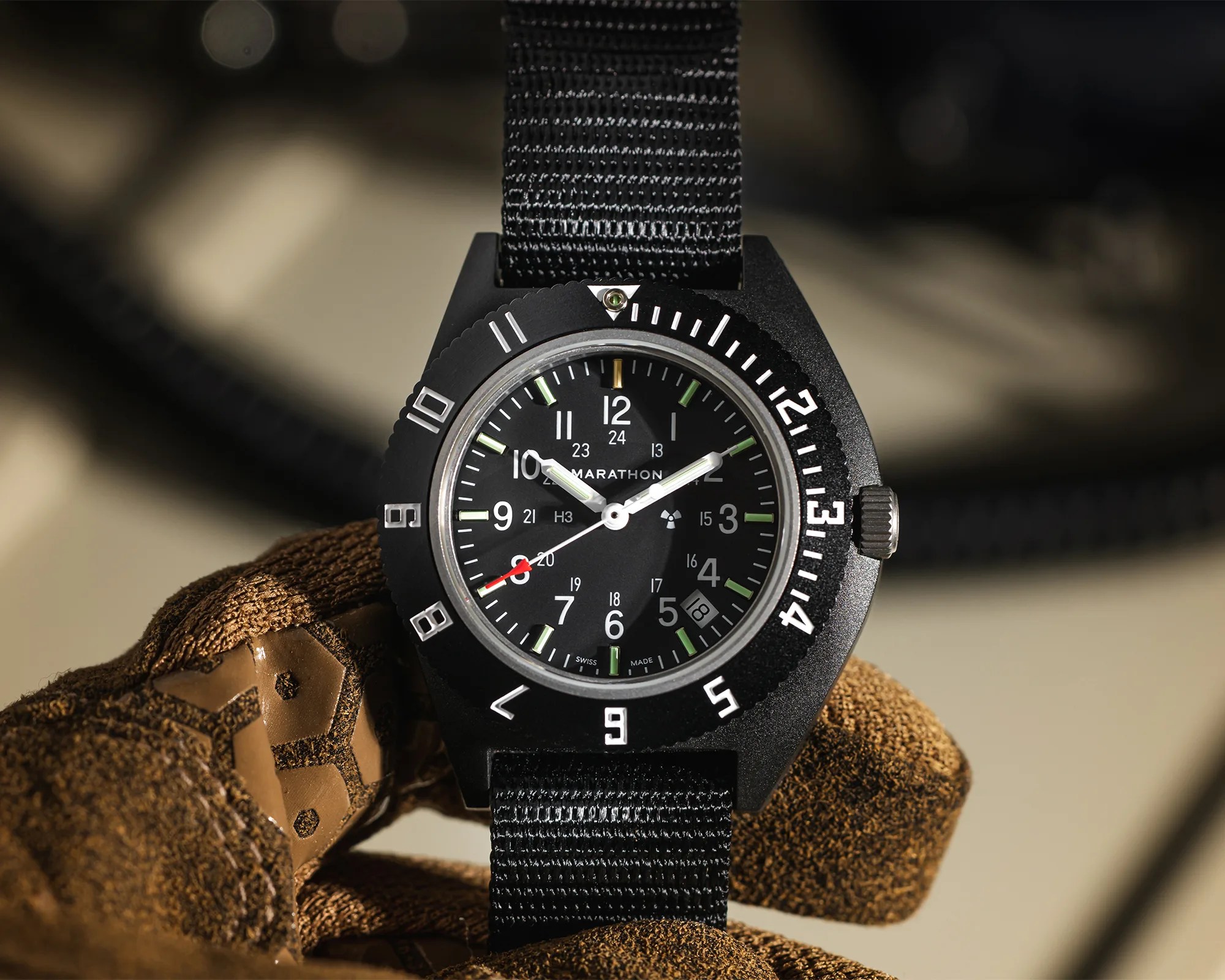 Black Marathon wristwatch with white numerals and green hour markers on a black nylon strap held in a brown glove.