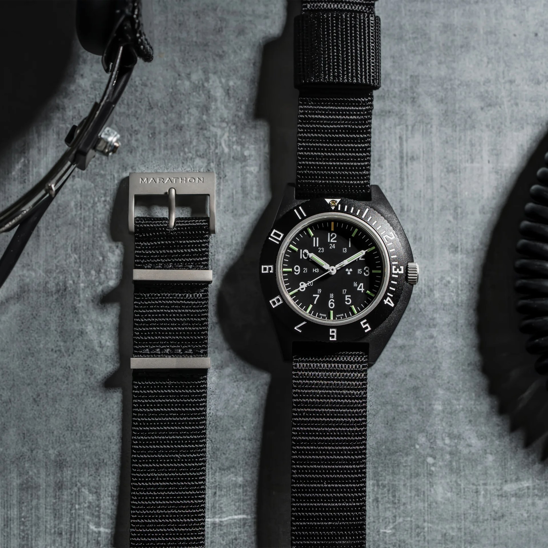 Black Marathon wristwatch with black nylon strap and silver buckle on a gray surface.
