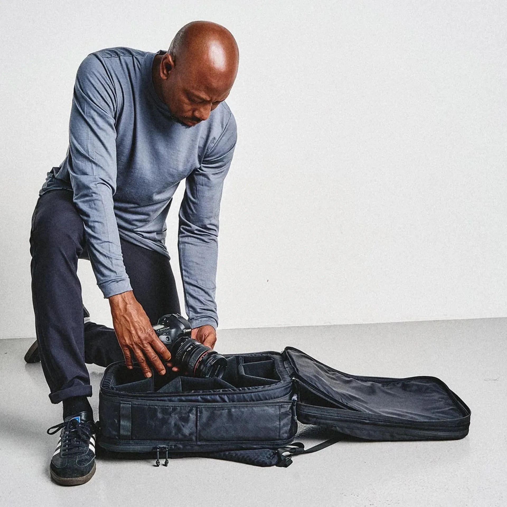 Man in blue long-sleeve shirt and black pants placing a Canon camera into a black padded camera bag on the floor.