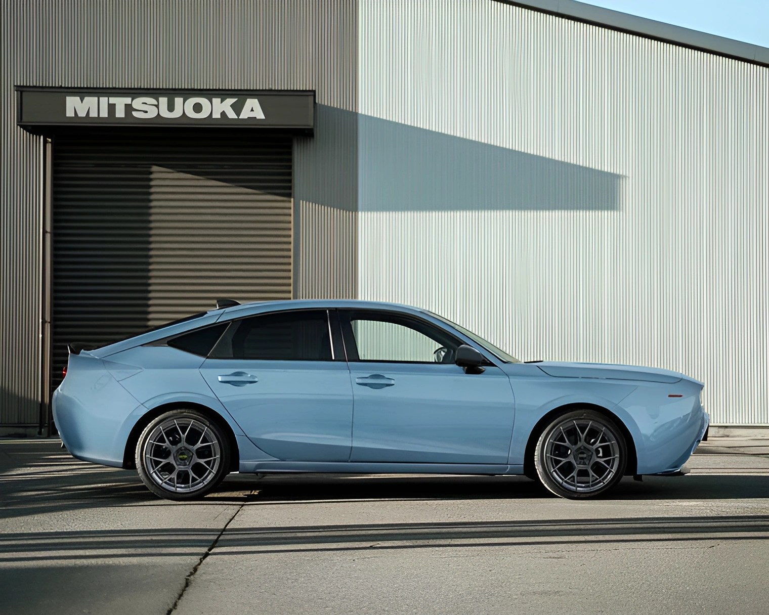 Light blue four-door sedan with sporty wheels parked in front of a building with a "MITSUOKA" sign.