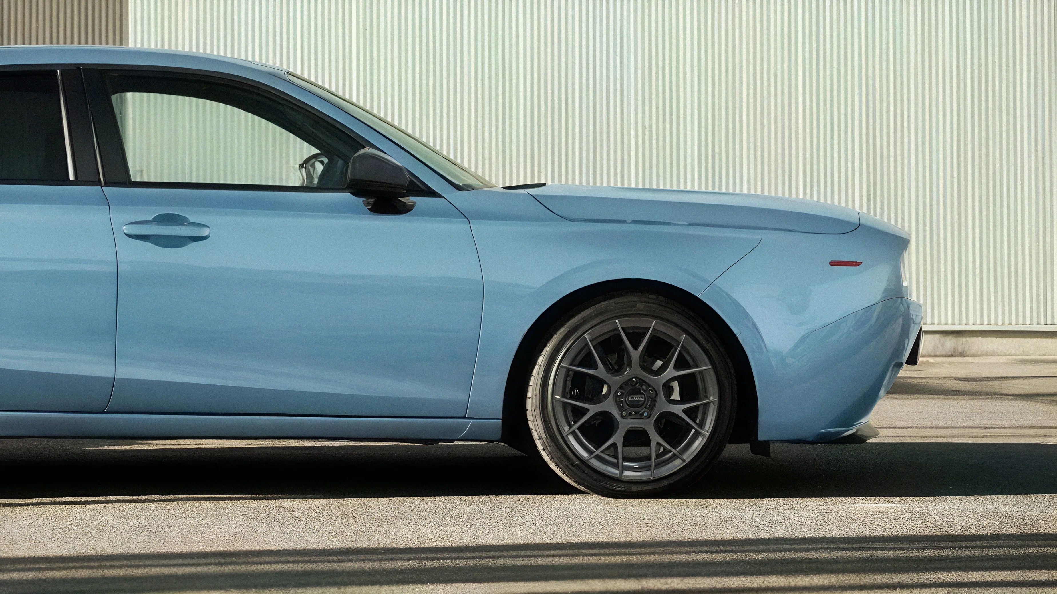 Light blue car side with dark gray multi-spoke alloy wheel and black side mirror against a corrugated wall.