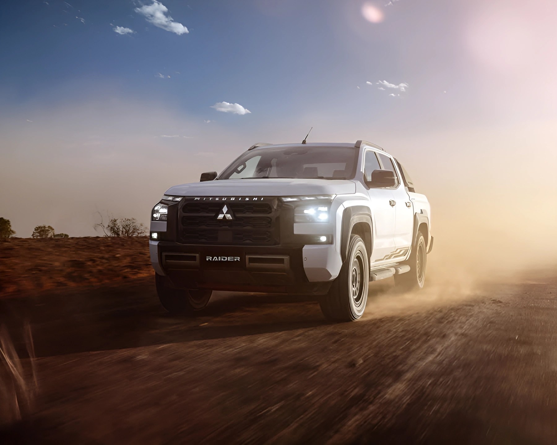 White Mitsubishi Raider pickup truck driving on a dusty dirt road at sunset.
