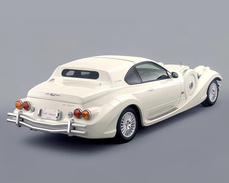 Cream-colored vintage-style luxury coupe with rounded fenders and chrome bumpers on a gray background.
