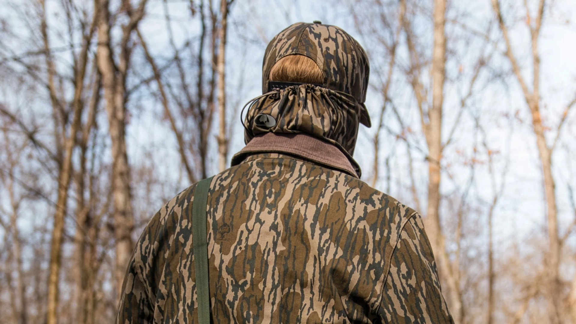 Person wearing a brown and green camouflage jacket and matching cap with a drawstring neck gaiter in a forest.