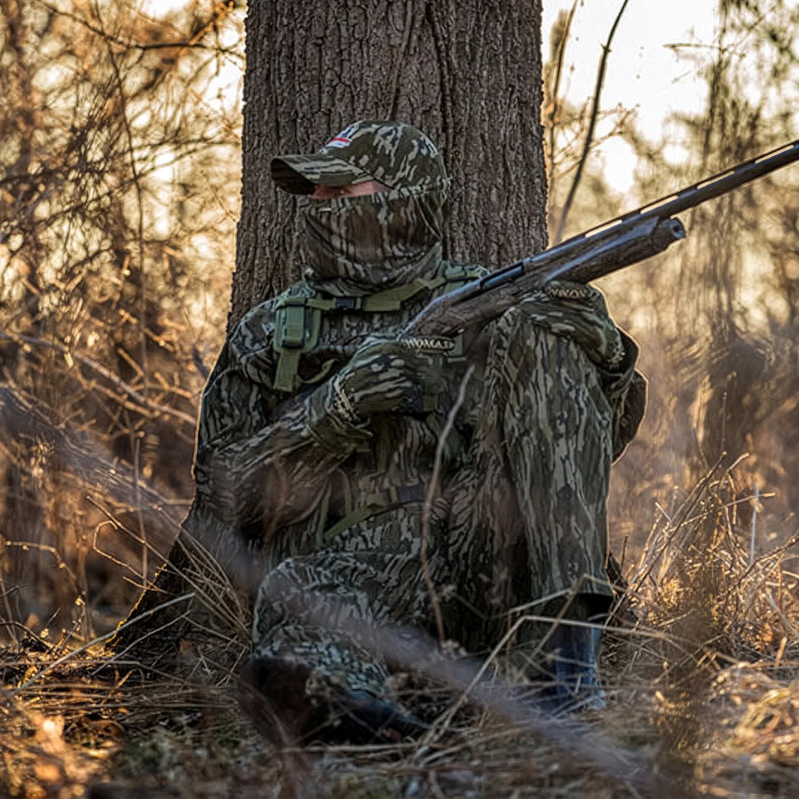 Person in full camouflage gear and face covering sitting against a tree holding a shotgun in a wooded area.