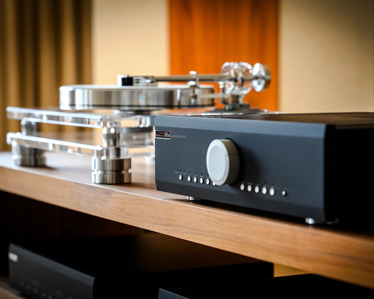 Black audio amplifier with a large silver knob and buttons on a wooden shelf next to a clear acrylic turntable.