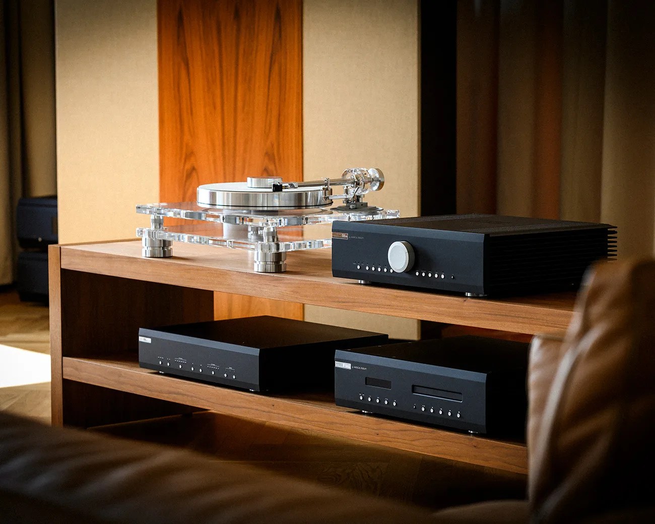 Clear acrylic turntable with silver platter and tonearm on a wooden shelf next to black audio components.