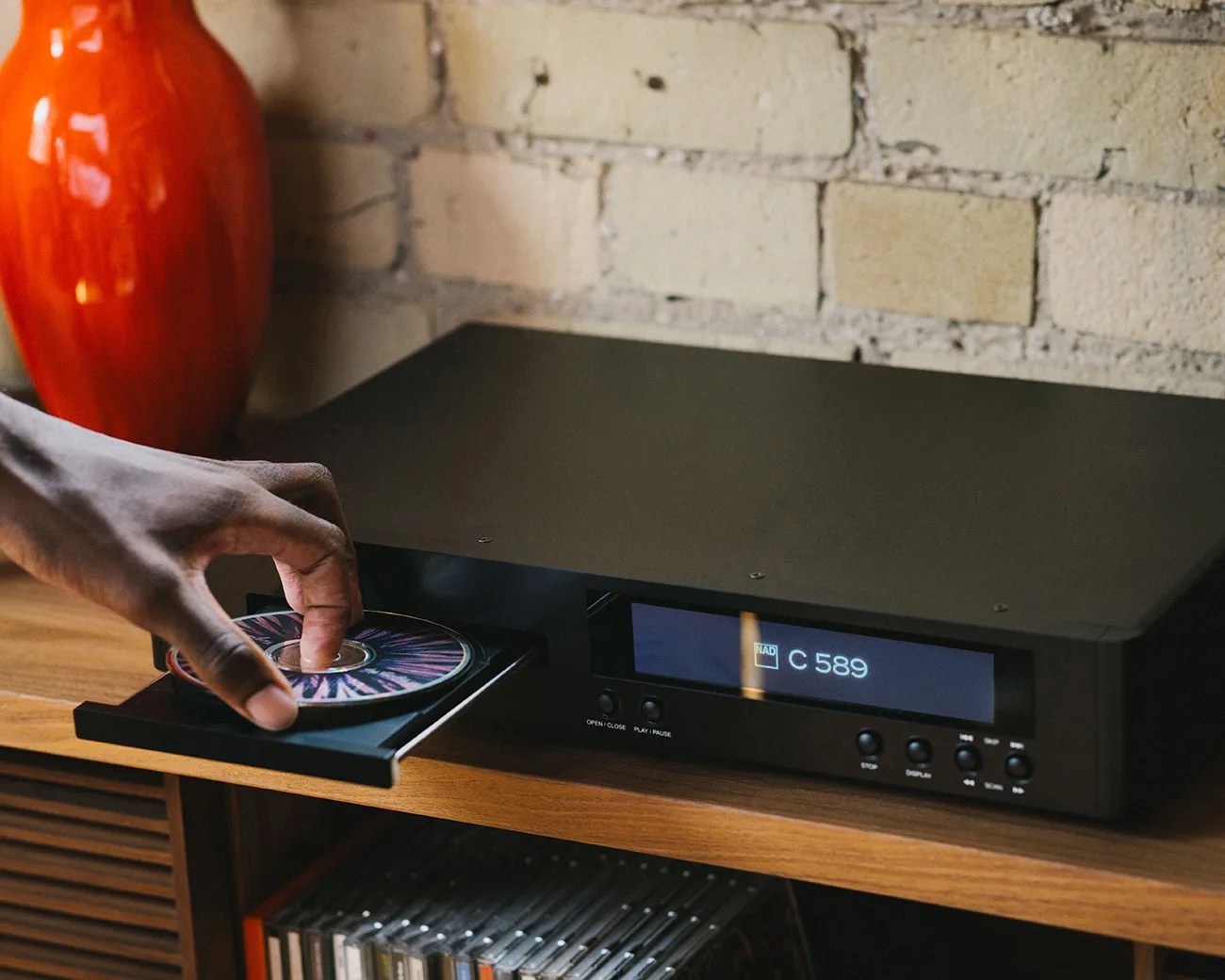 Hand placing a CD with a colorful design into a black NAD C 589 CD player on a wooden shelf.