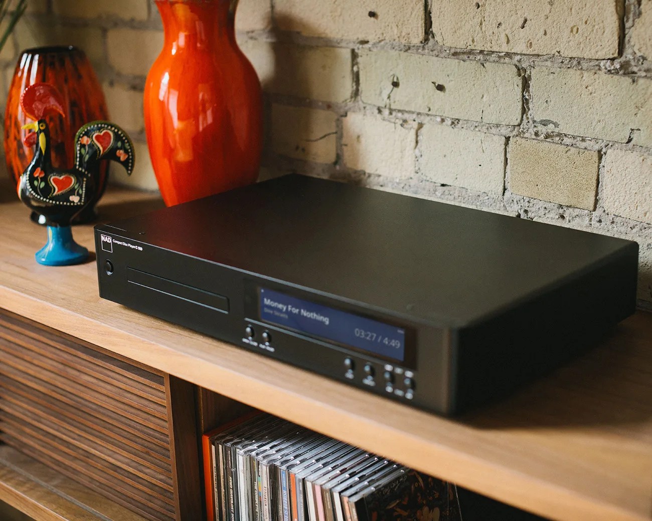 Black NAD Compact Disc Player C 576 with digital display showing "Money For Nothing" on a wooden shelf with CDs below and decorative items behind.