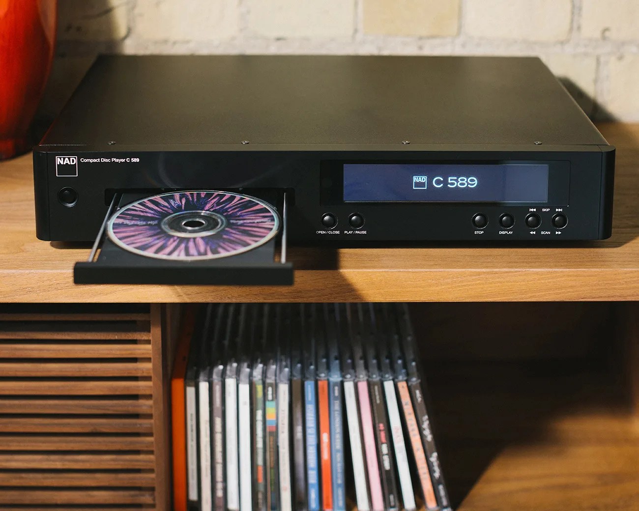 Black NAD C 589 compact disc player with tray open holding a CD, placed on a wooden shelf above a row of CDs.