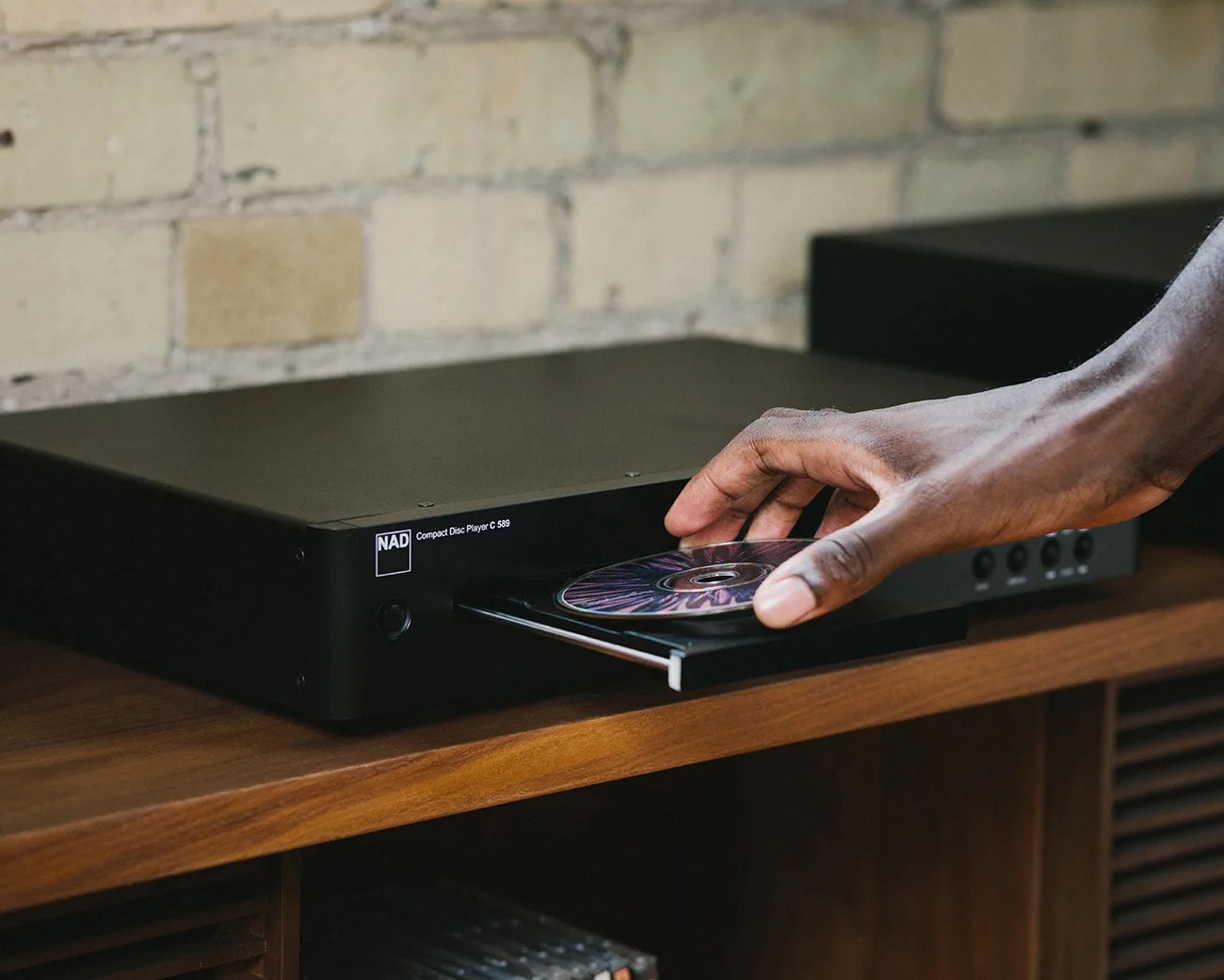Hand placing a CD into the tray of a black NAD Compact Disc Player C 589 on a wooden shelf.