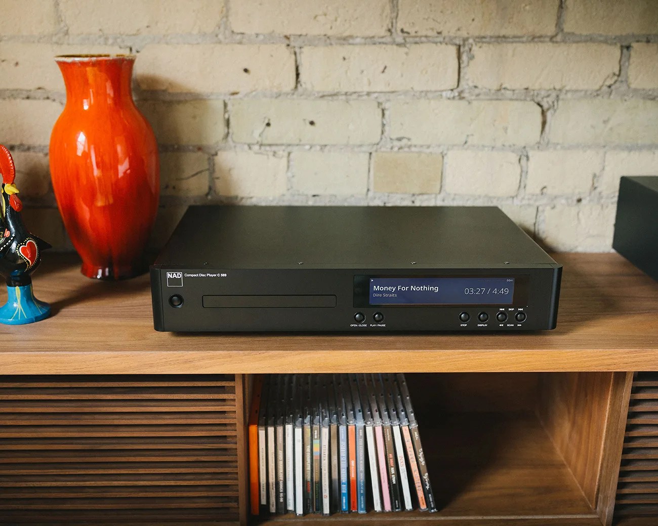 Black NAD Compact Disc Player C 589 on a wooden shelf playing "Money For Nothing" by Dire Straits.