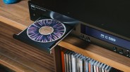 Black NAD C 589 compact disc player with an open tray holding a CD with a purple and black abstract design, placed on a wooden shelf above a collection of CDs.