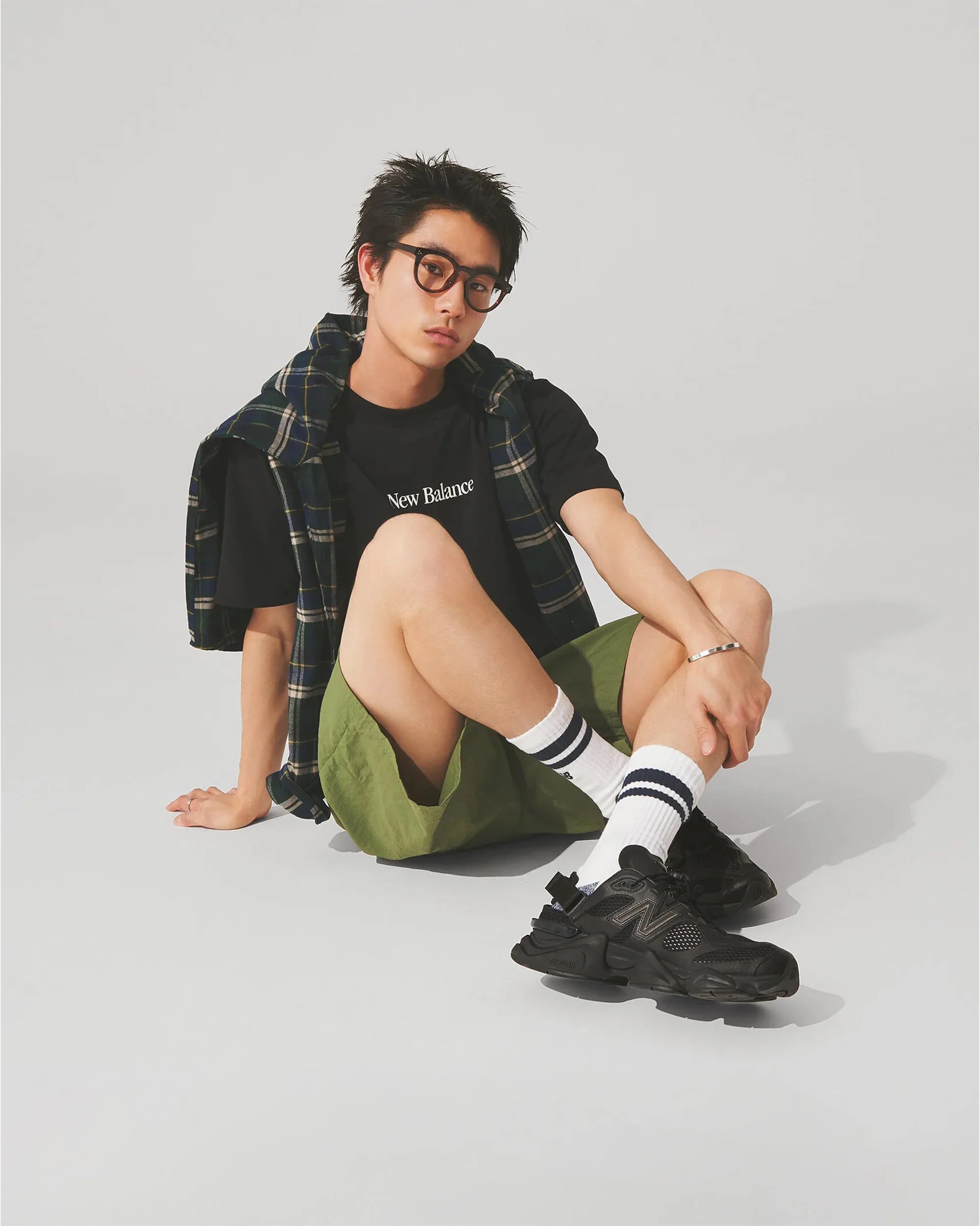 Person sitting on the floor wearing black New Balance t-shirt, green shorts, black sneakers, white socks with black stripes, and a plaid shirt draped over shoulders.