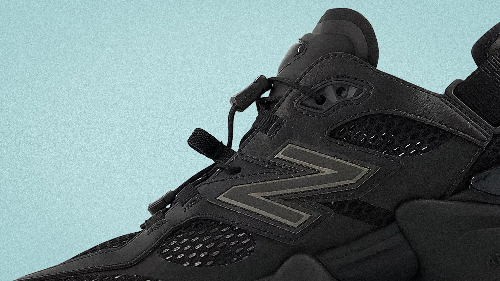 Close-up of a black New Balance sneaker with mesh panels and a reflective "N" logo on a light blue background.