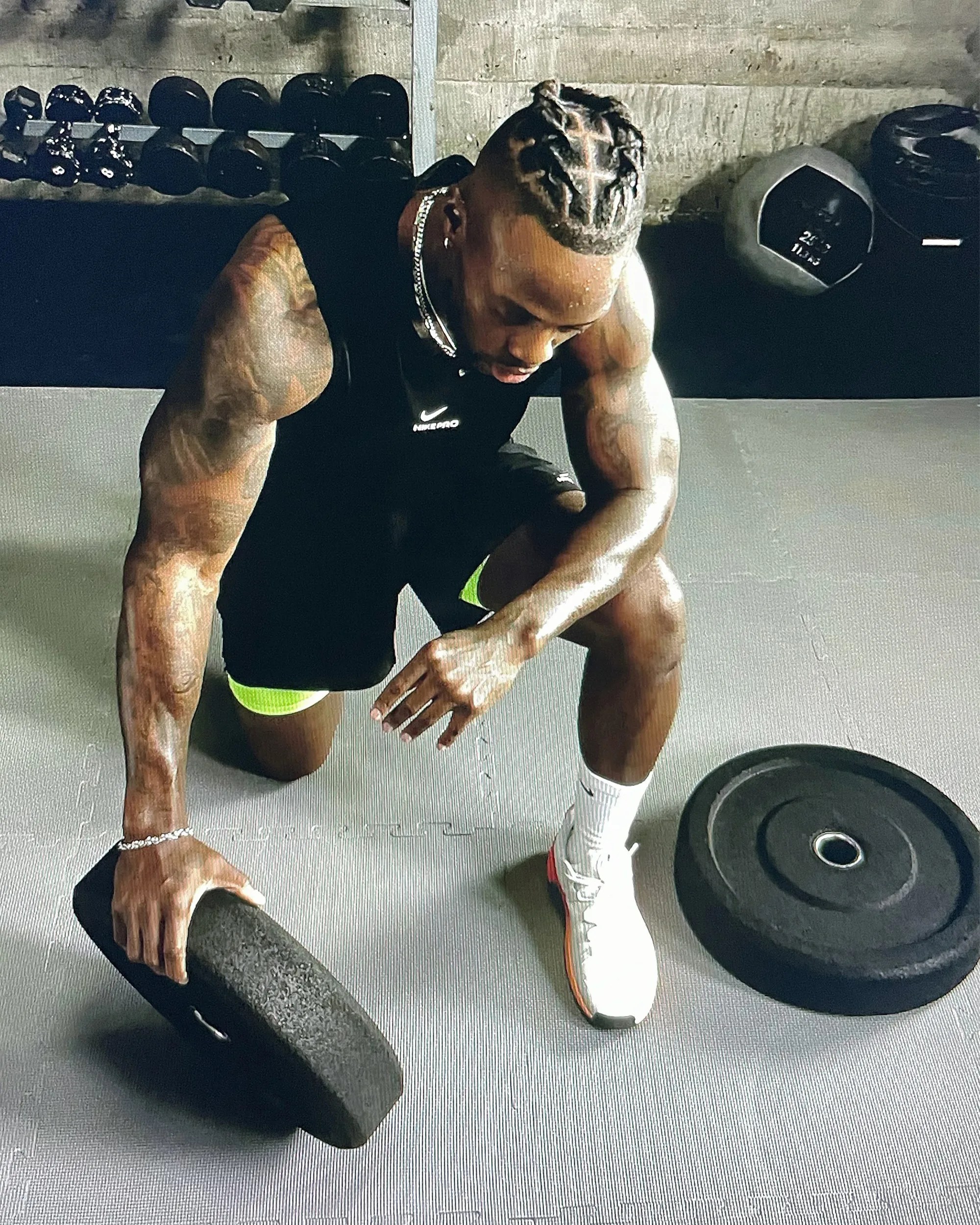 Muscular man with braided hair in black Nike tank and shorts kneeling on gym floor holding a weight plate.