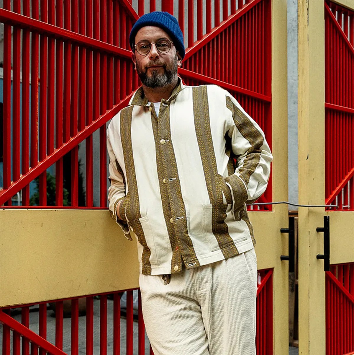Man wearing a cream jacket with brown vertical stripes and cream pants, standing against a red metal gate.