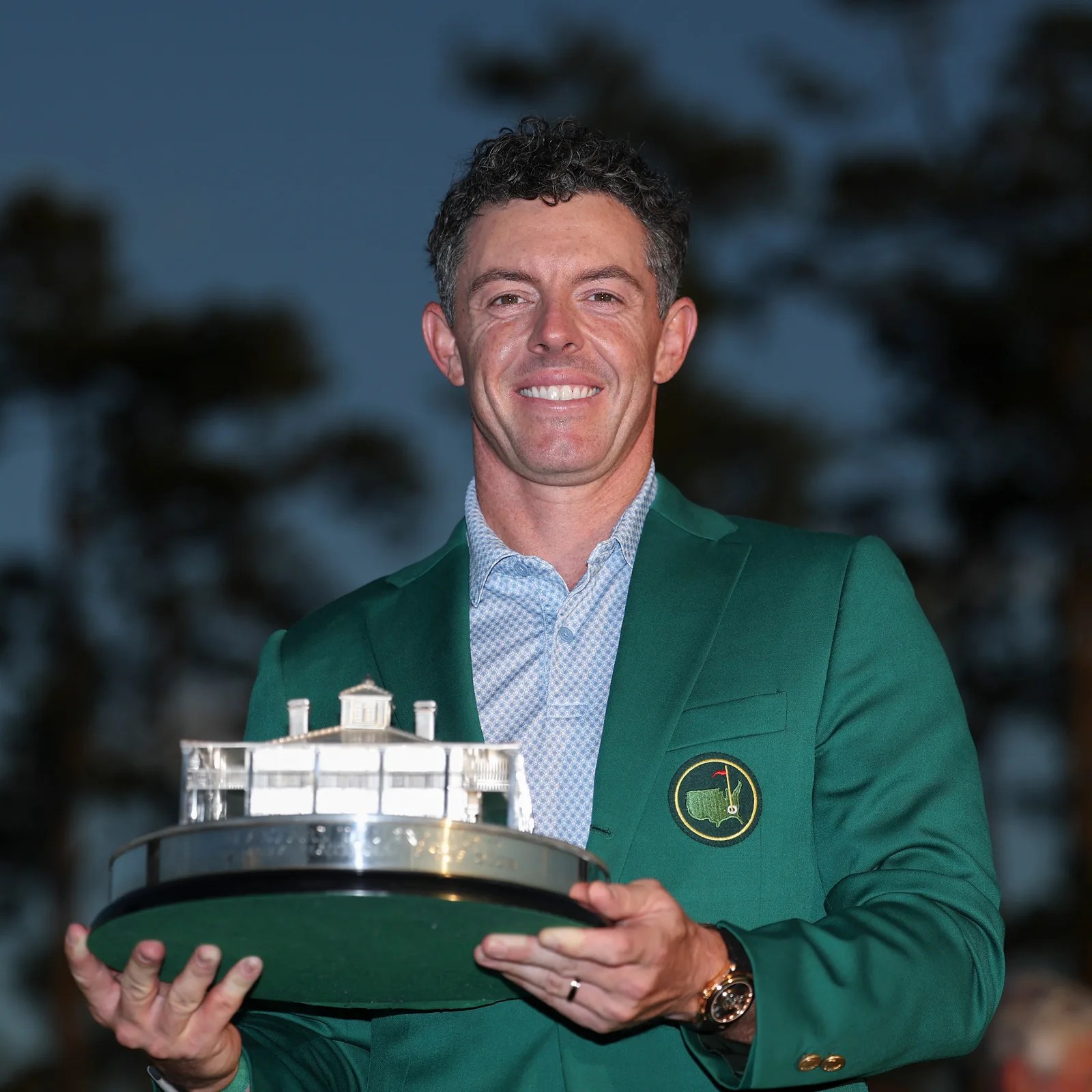 Man wearing a green Masters jacket holding a silver clubhouse-shaped trophy against a dark outdoor background.