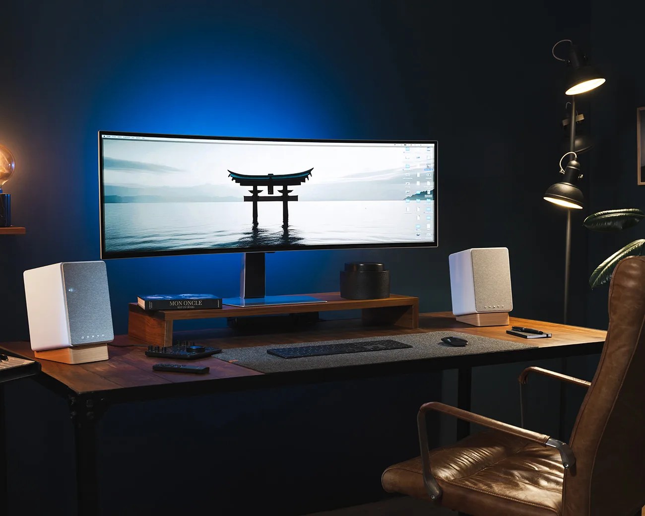 Wooden desk setup with an ultrawide curved monitor displaying a torii gate, white speakers on wooden stands, and a brown leather chair.