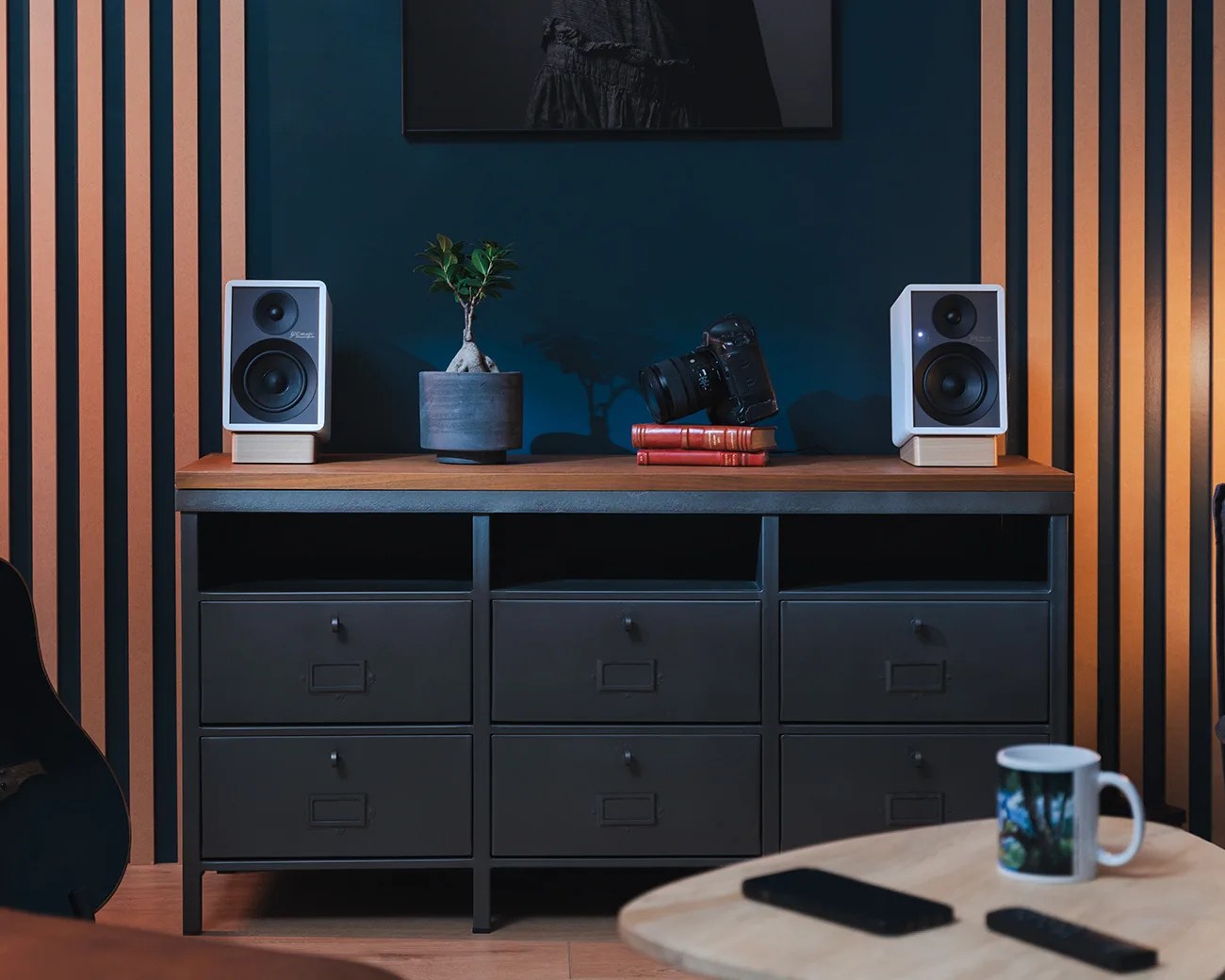 Black metal cabinet with six drawers topped with wooden surface holding two white speakers, a potted plant, and a camera on books.