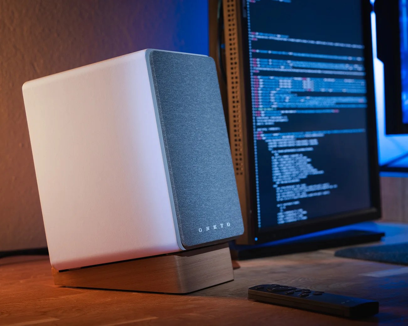 White Onkyo speaker with gray fabric front on a wooden stand beside a computer monitor and remote on a desk.