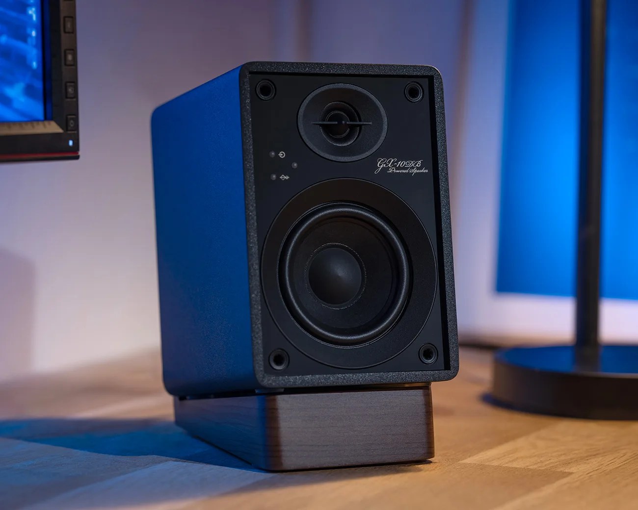 Black GX-10DB powered speaker with a blue casing on a wooden stand on a desk.