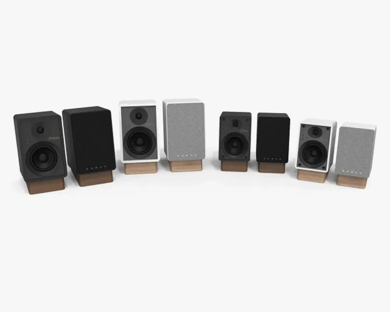 Four pairs of compact rectangular speakers in black and white finishes, each pair with one speaker showing drivers and the other covered with a fabric grille, all on wooden stands.