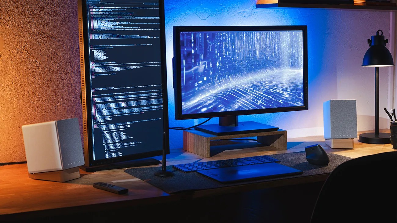 Dual computer monitors on a wooden desk with vertical coding screen, horizontal display with blue digital graphics, and gray speakers.