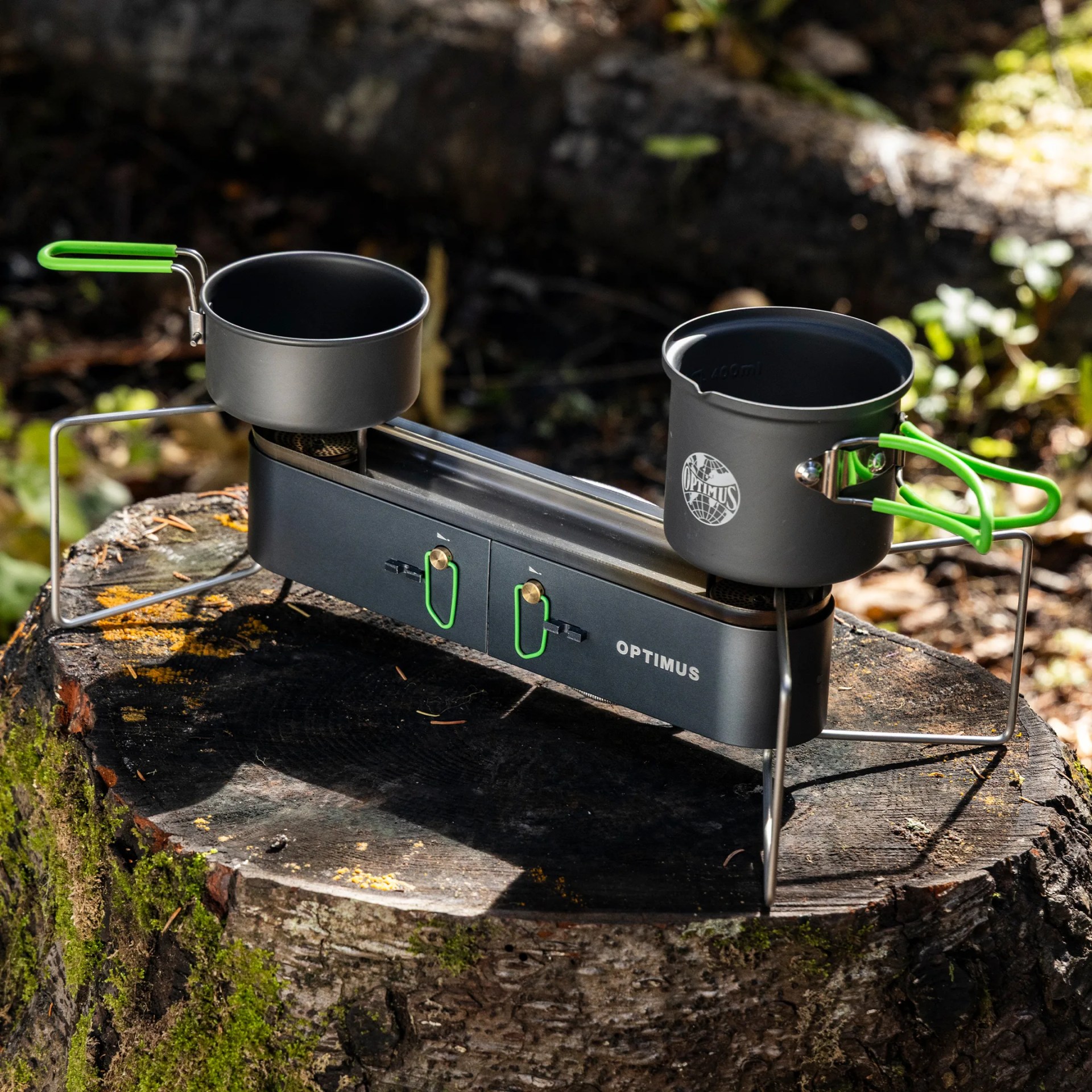 Black portable camping stove with two burners and green-handled pots on a tree stump outdoors.