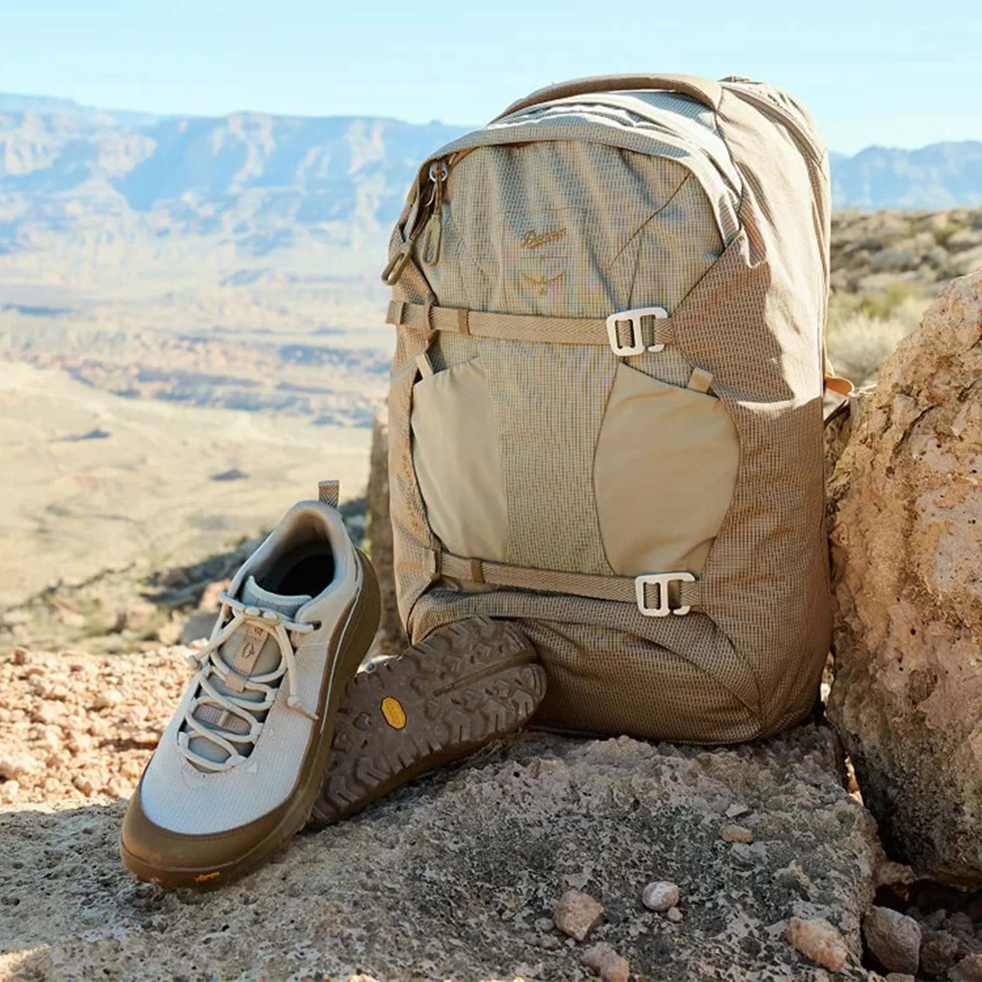 Tan and beige hiking backpack with buckles next to a pair of beige and brown hiking shoes on rocky terrain.