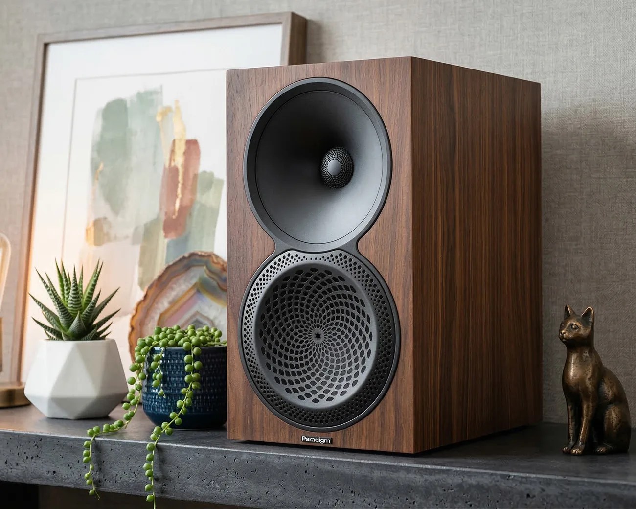 Wood-finished Paradigm speaker with black circular drivers on a concrete shelf next to plants and a cat figurine.