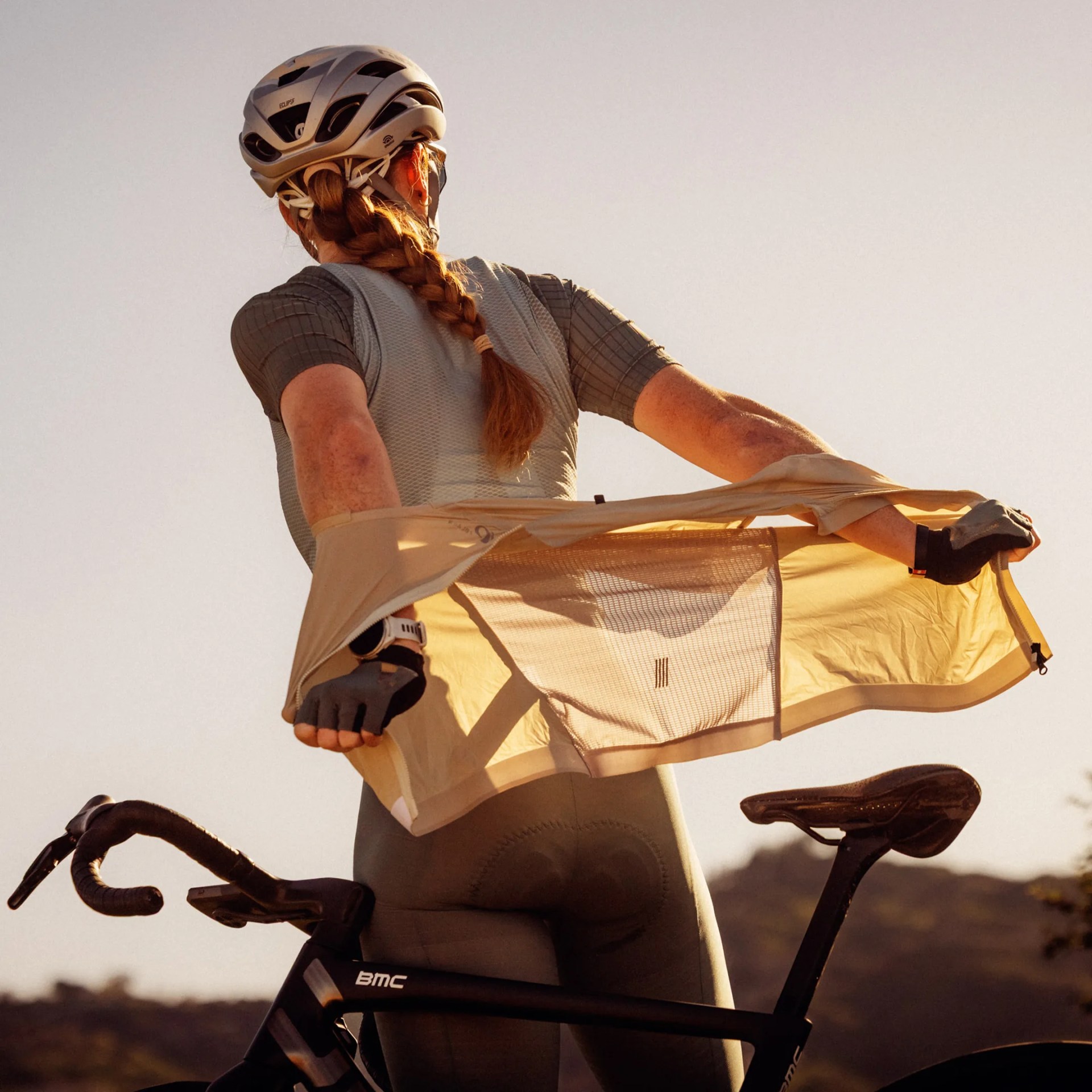 Cyclist wearing a gray helmet and gloves putting on a light tan cycling jacket while standing next to a black BMC road bike.