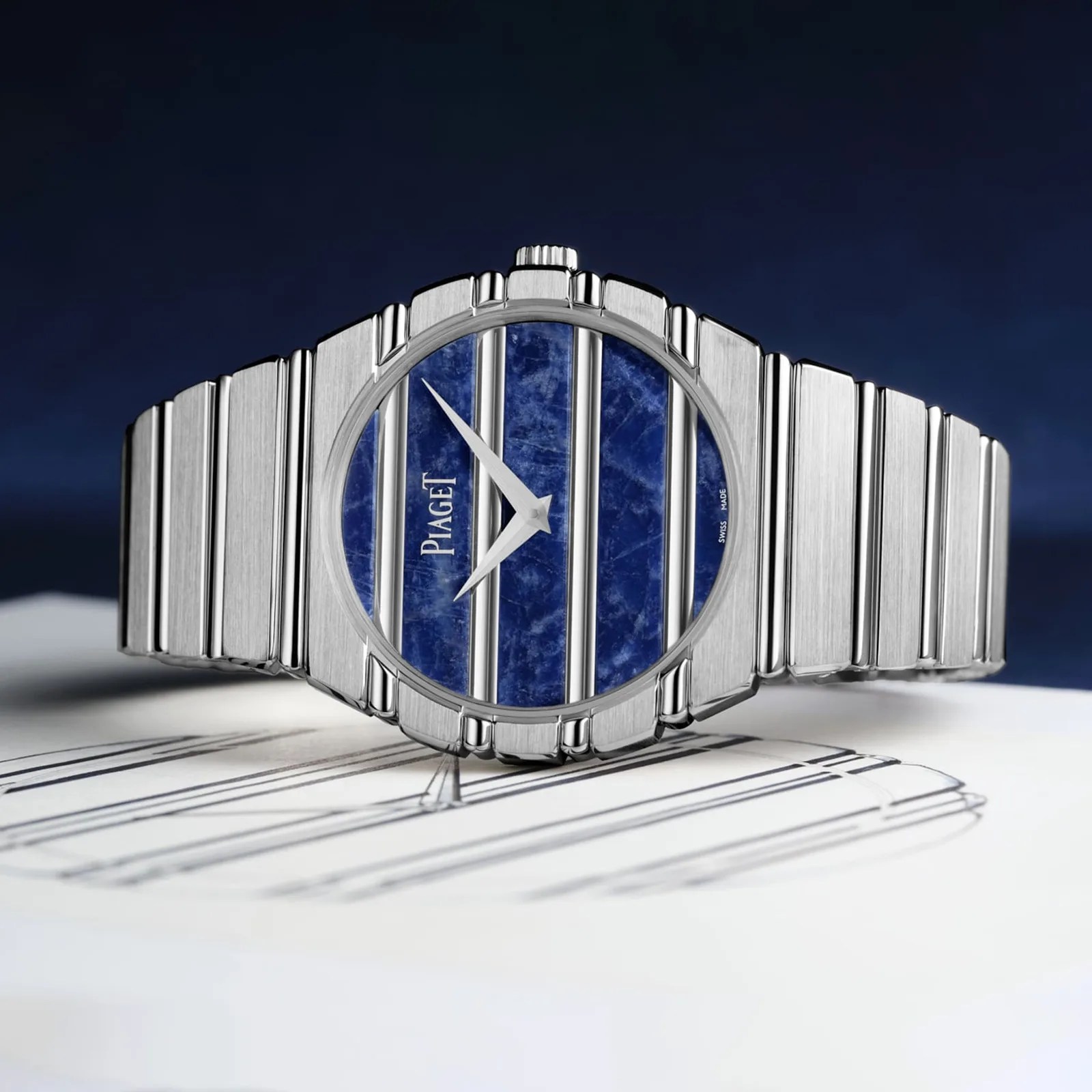 Silver metal wristwatch with a blue and silver striped dial and "Piaget" logo on a white surface.