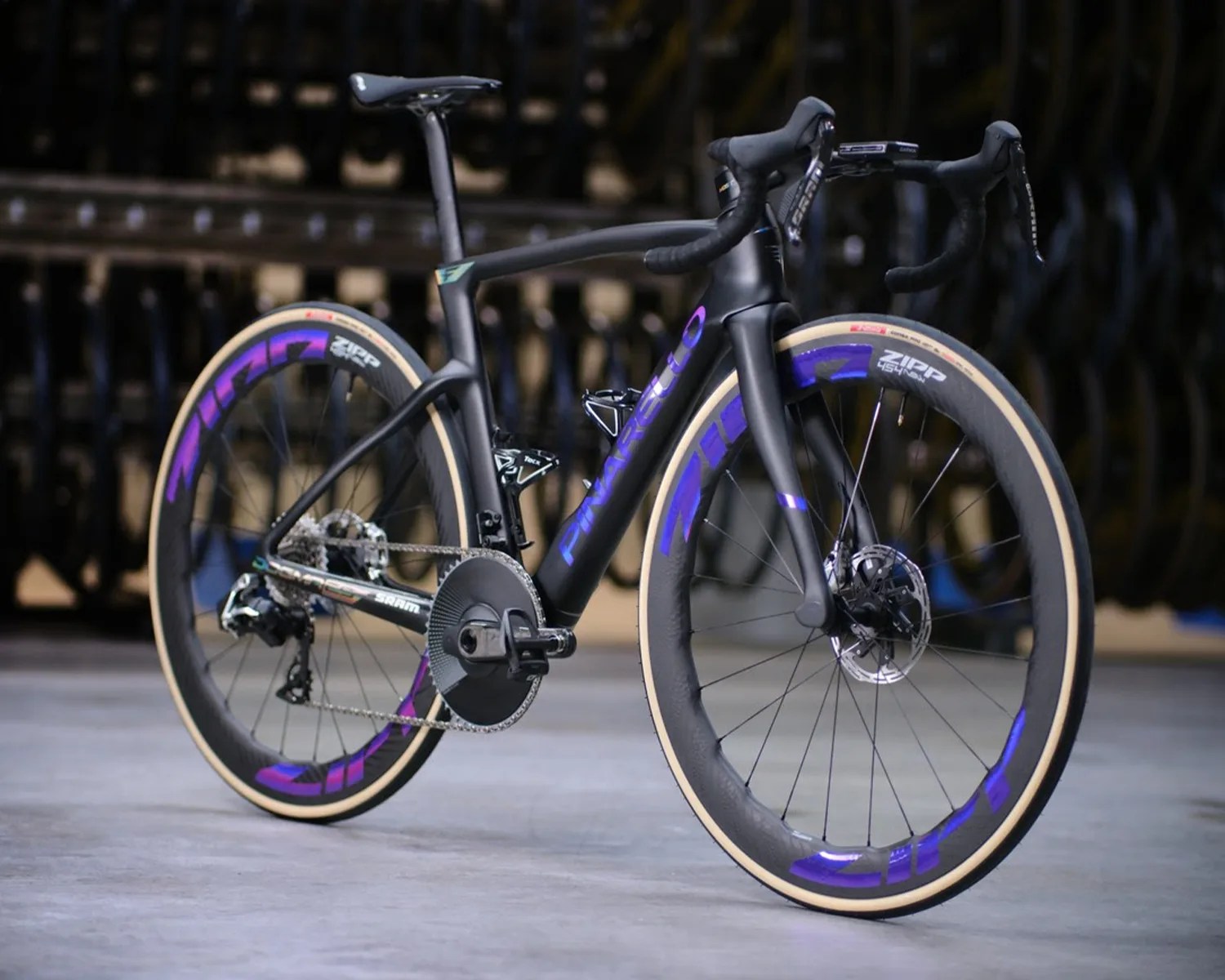 Matte black Pinarello road bike with purple Zipp 454 NSW wheels and tan sidewall tires on a concrete floor.