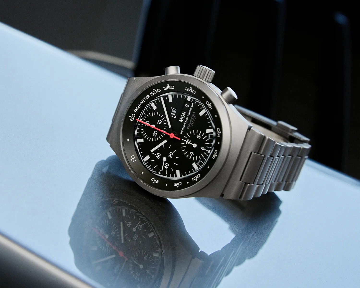 Matte silver chronograph wristwatch with black dial, white markers, red second hand, and metal link bracelet on reflective surface.