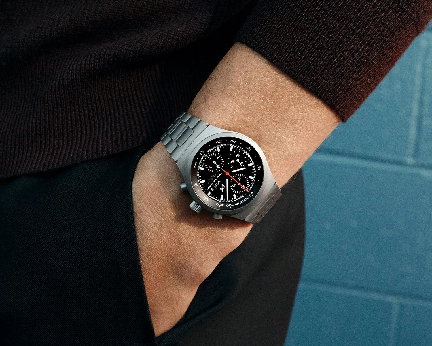 Silver metal chronograph wristwatch with black dial and red second hand worn on a wrist with a hand in a pocket.