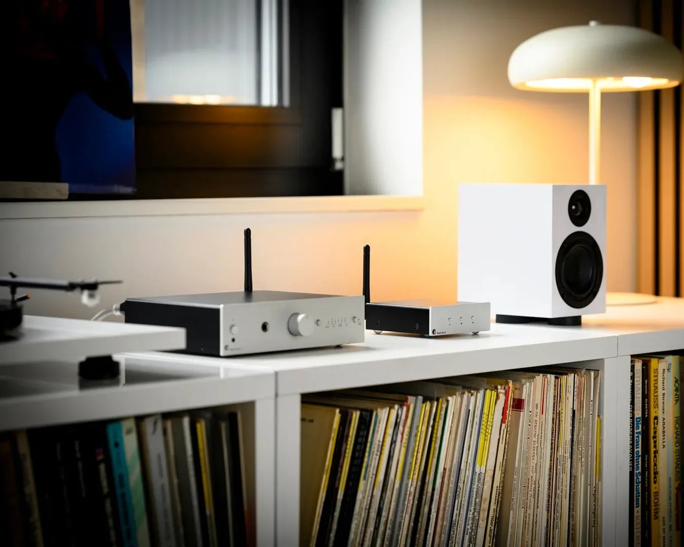 White bookshelf with vinyl records, a white speaker, silver audio equipment with antennas, and a white table lamp.