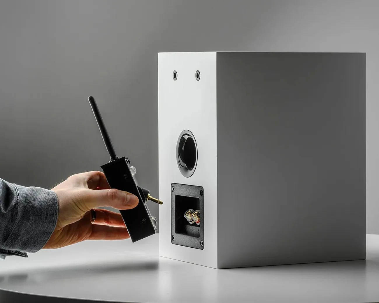 Hand holding a black wireless audio transmitter near the back panel of a white speaker on a gray surface.