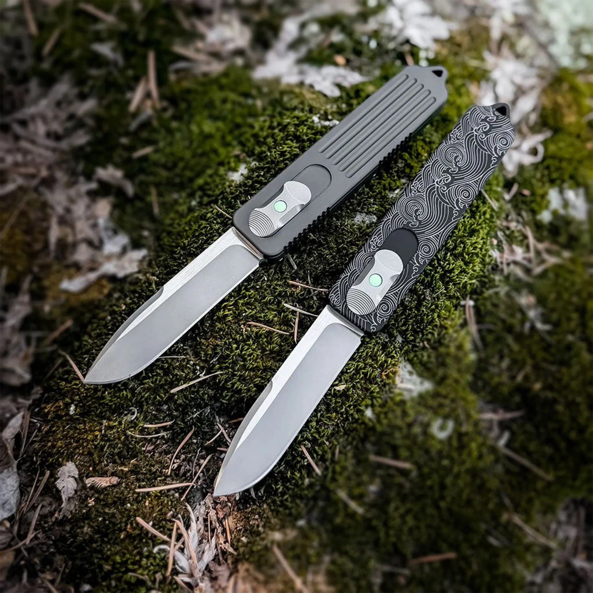 Two folding knives with silver blades and different textured handles, one plain gray and one black with white wave patterns, placed on green moss.