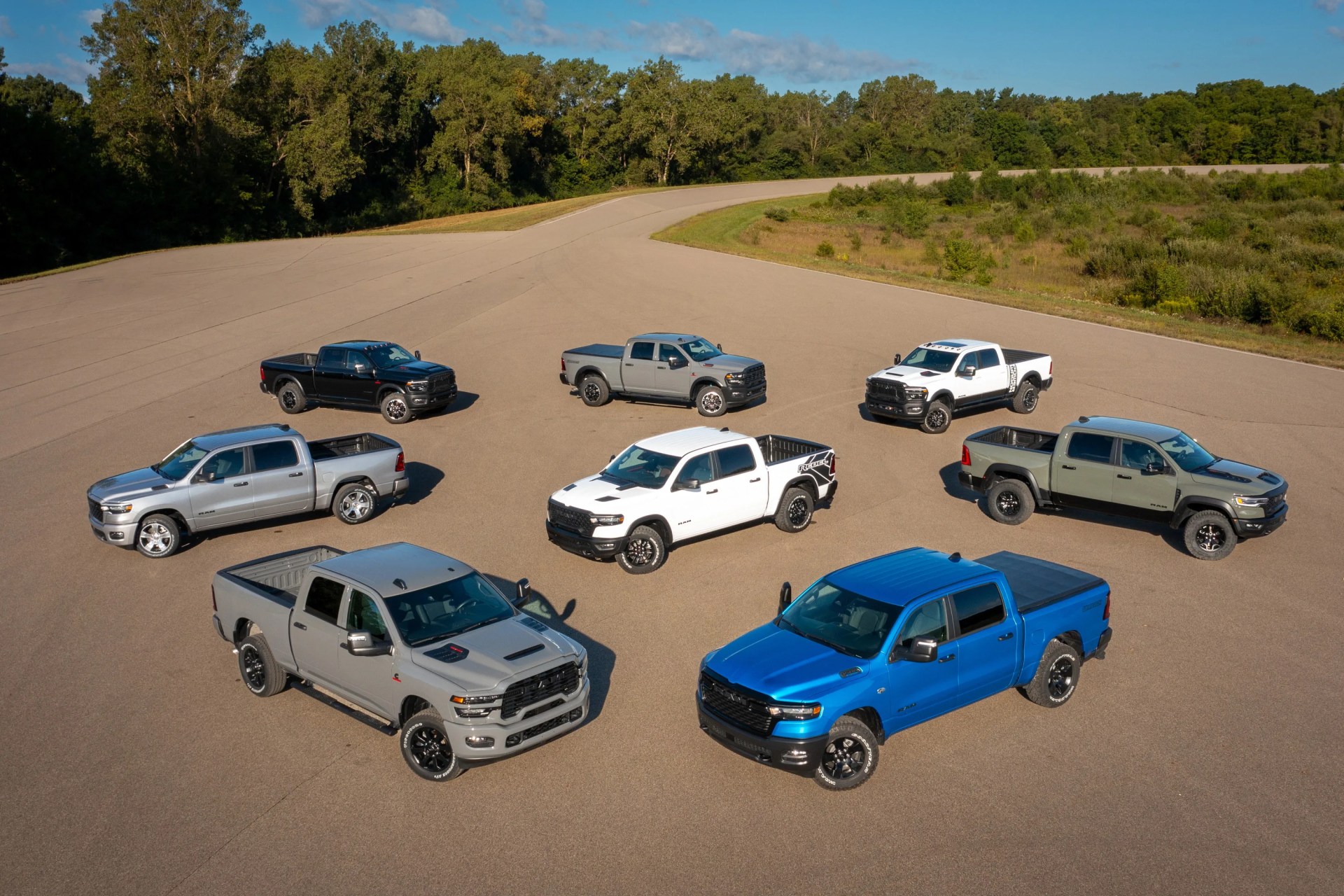 Eight pickup trucks in various colors, including blue, white, gray, black, and silver, arranged in a circle on a paved open area with trees in the background.