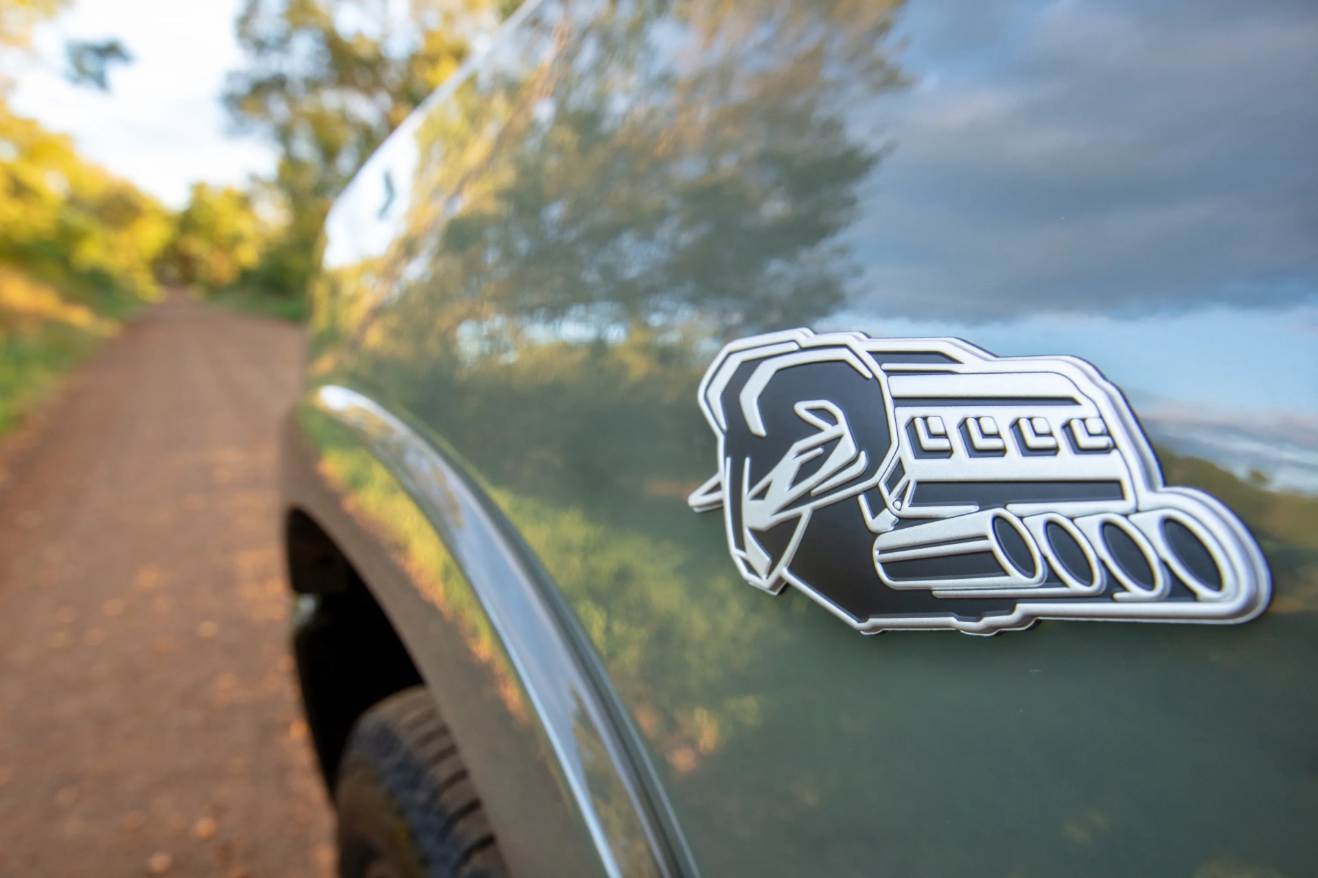 Close-up of a silver and black engine emblem on the side of a green vehicle with a dirt road and trees reflected on the surface.