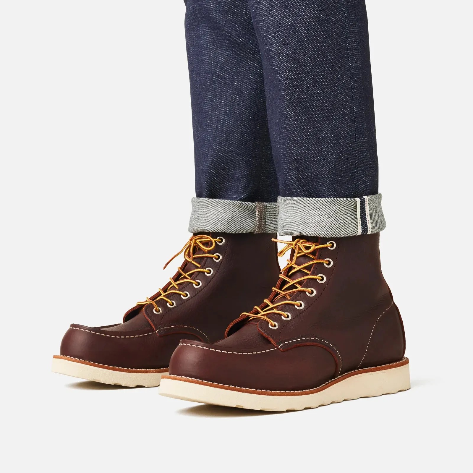 Brown leather moc toe boots with yellow laces and white soles worn with cuffed dark blue denim jeans.
