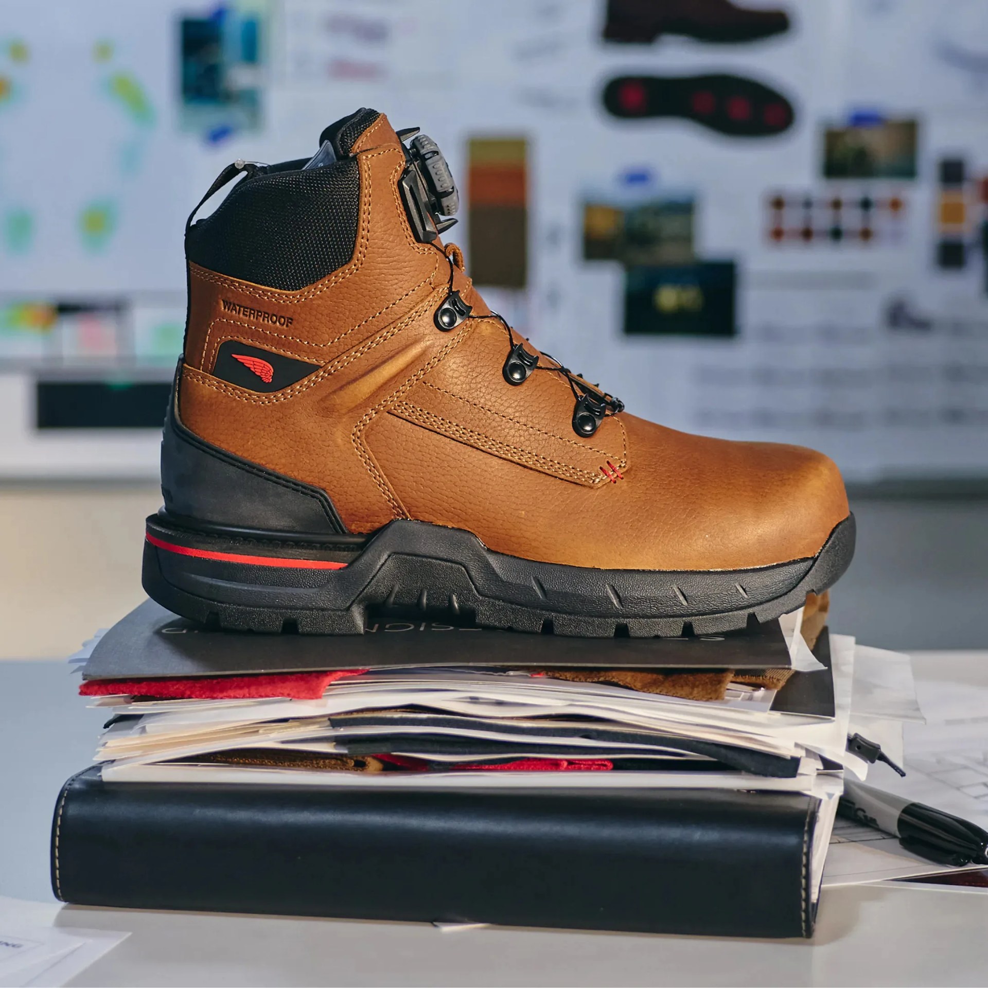 Brown leather waterproof hiking boot with black sole and red wing logo on a stack of papers.