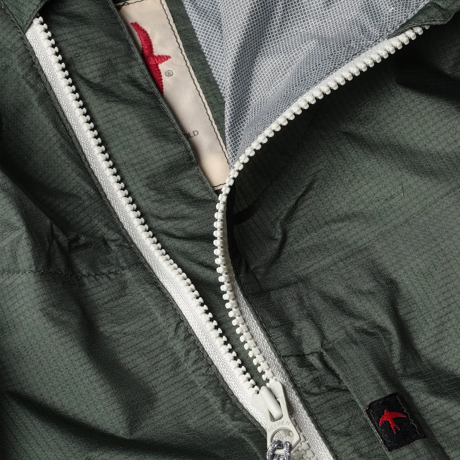 Close-up of a dark green jacket with a white zipper partially open, showing a gray mesh lining and a red bird logo on a black tag.