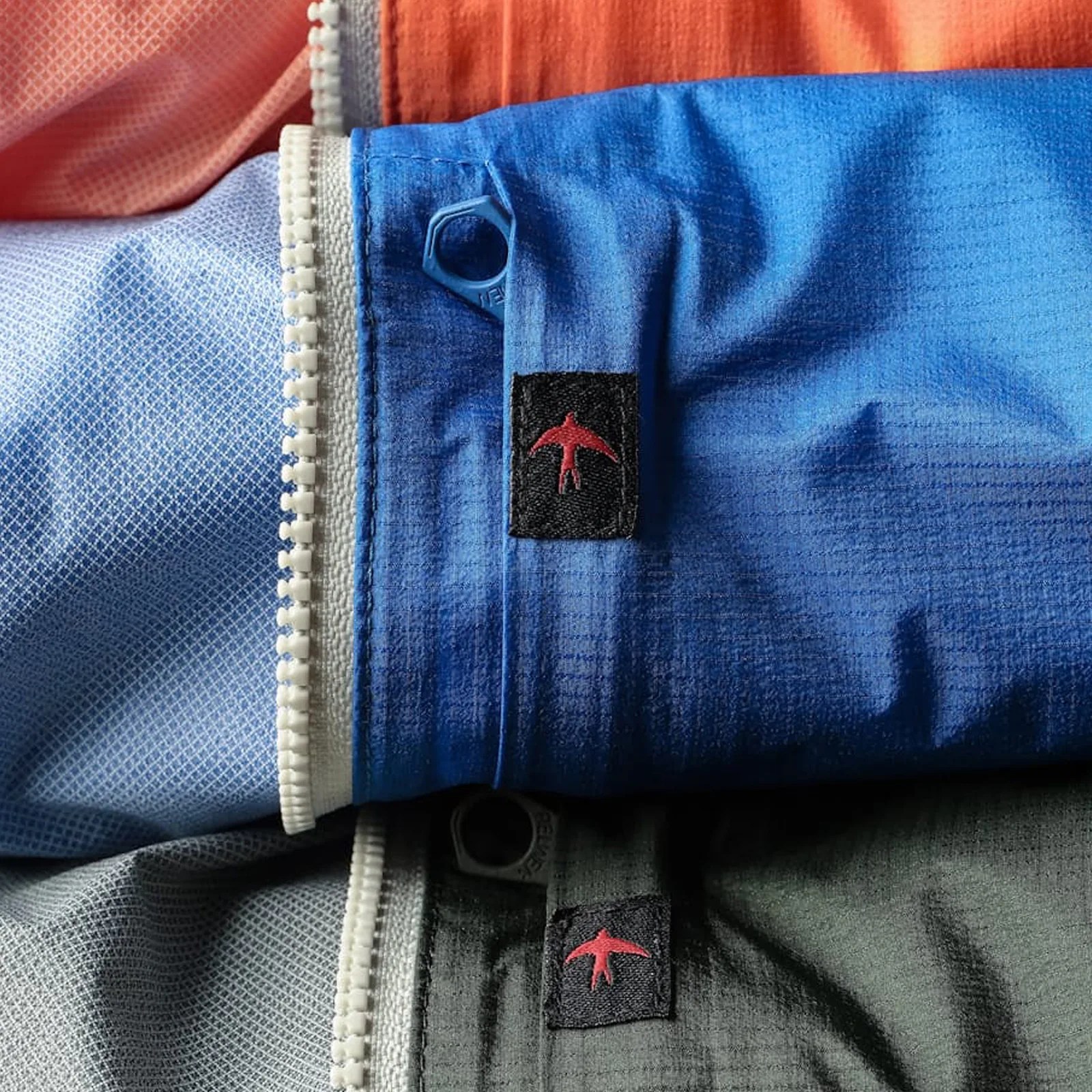 Close-up of blue and gray fabric with white zipper and small black tags featuring a red bird logo.