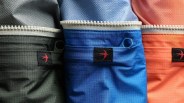 Close-up of three colorful fabric bags with white zippers and small black patches featuring a red bird logo.