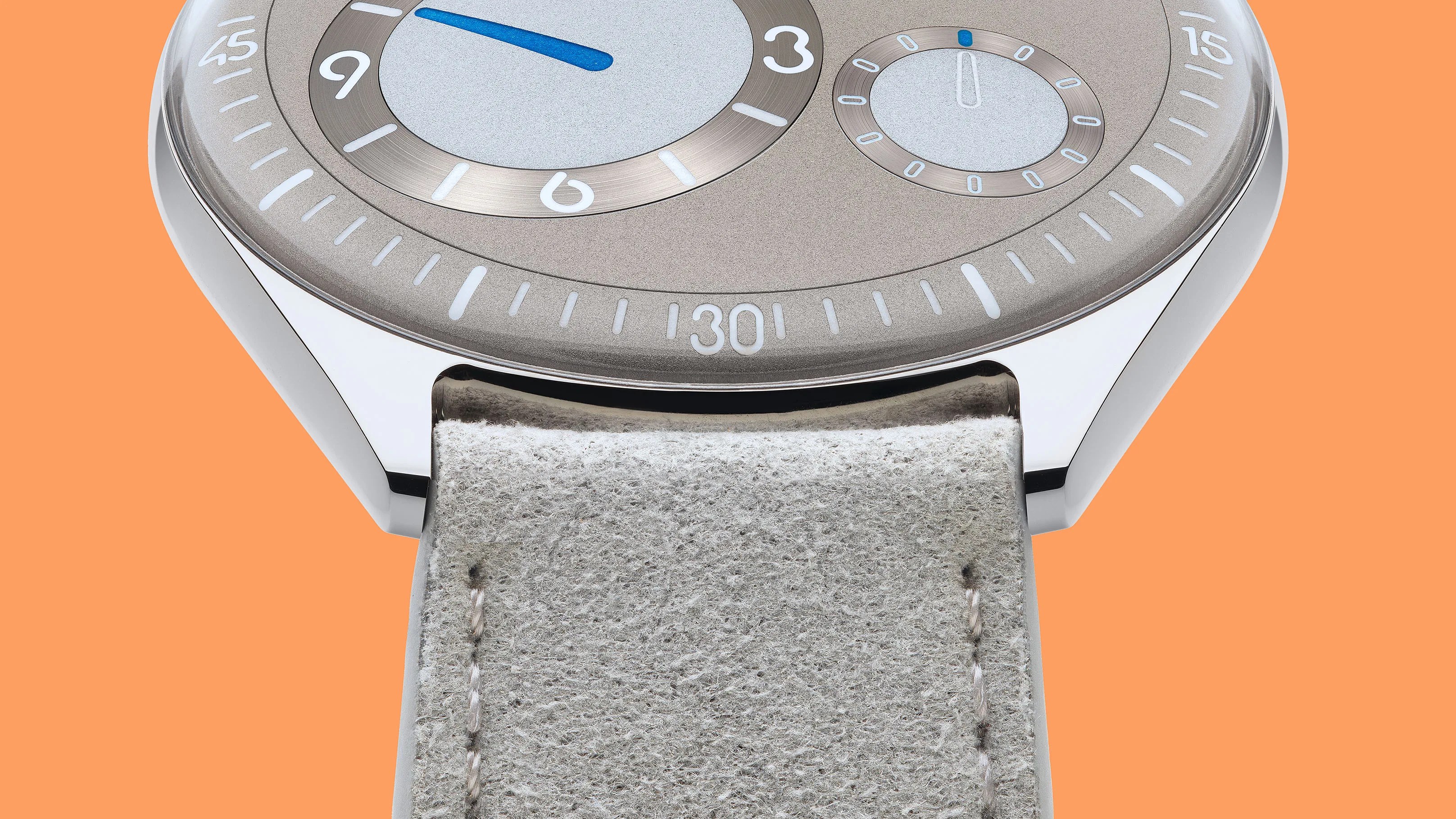 Close-up of a watch with a polished silver case, taupe dial, blue hand, and light gray suede strap against an orange background.