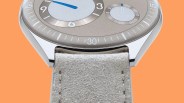 Close-up of a watch with a polished silver case, taupe dial, blue hand, and light gray suede strap against an orange background.